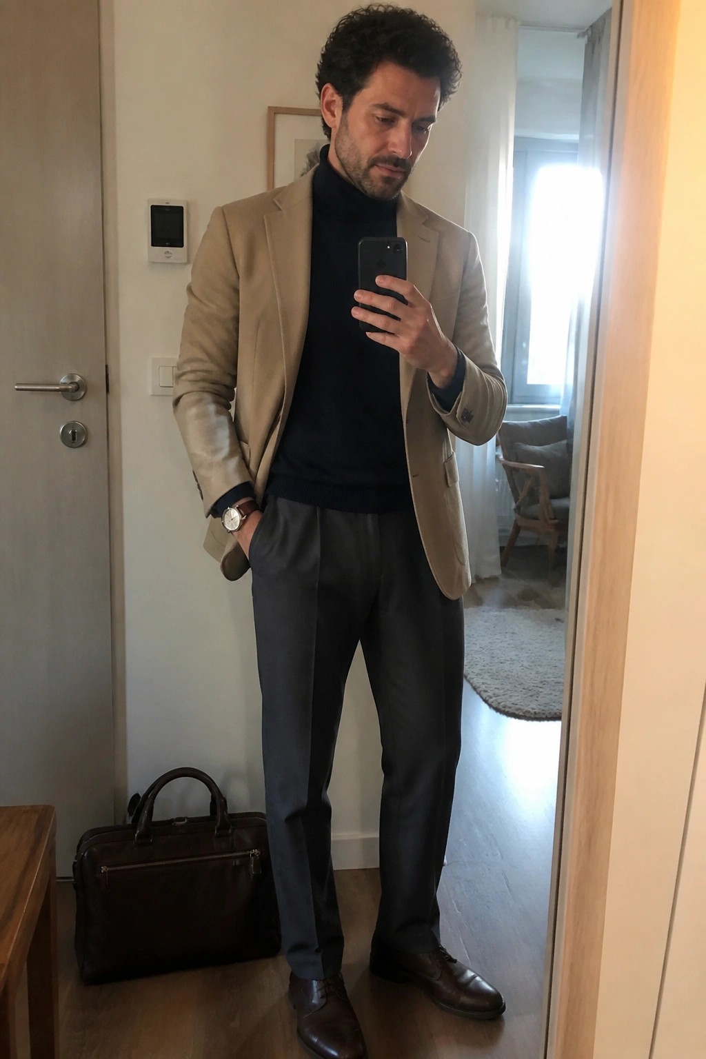Man in beige blazer over navy turtleneck sweater with grey trousers brown leather shoes and brown watch holding a phone next to a leather bag.