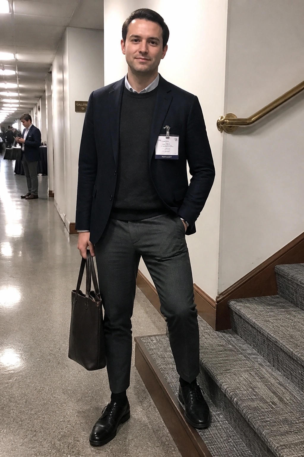 Man wearing navy blazer over dark gray sweater and light shirt, paired with gray trousers and black shoes while holding a brown leather tote on indoor stairs.