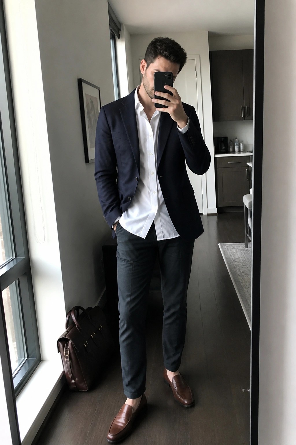 Man in navy blazer and white shirt taking mirror selfie in modern apartment