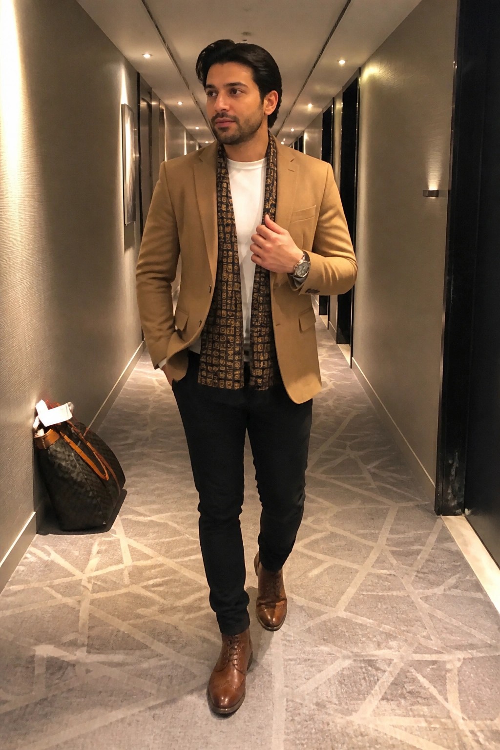 Man in tan blazer and patterned scarf stands in modern hotel hallway.