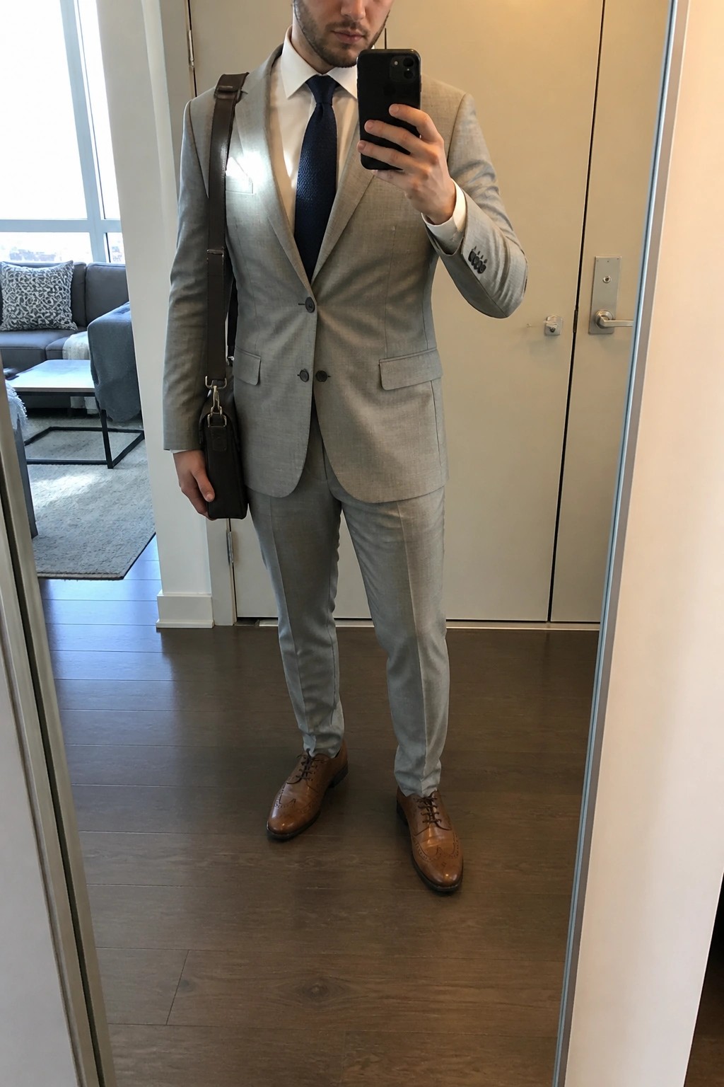 Man in light gray suit taking mirror selfie with phone and brown bag