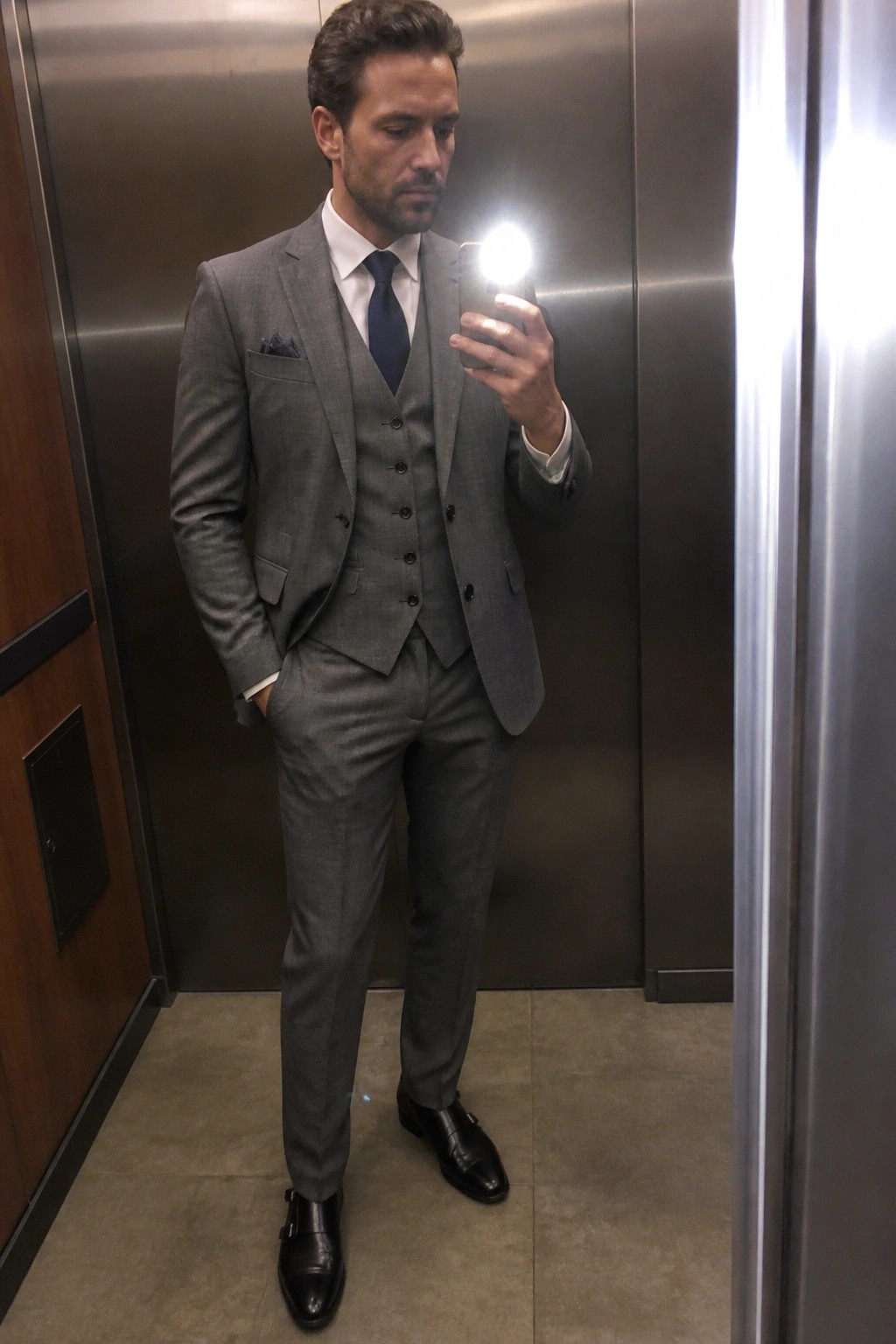 Man in gray three-piece suit taking mirror selfie in elevator.