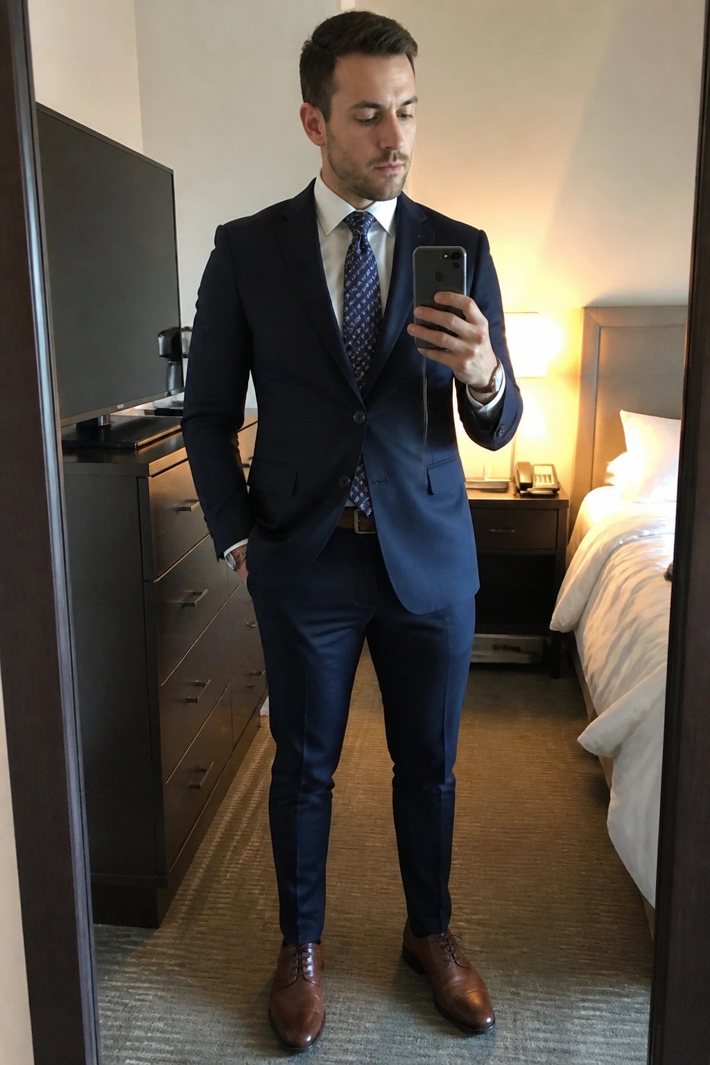 Man in navy suit taking mirror selfie with phone in hotel room