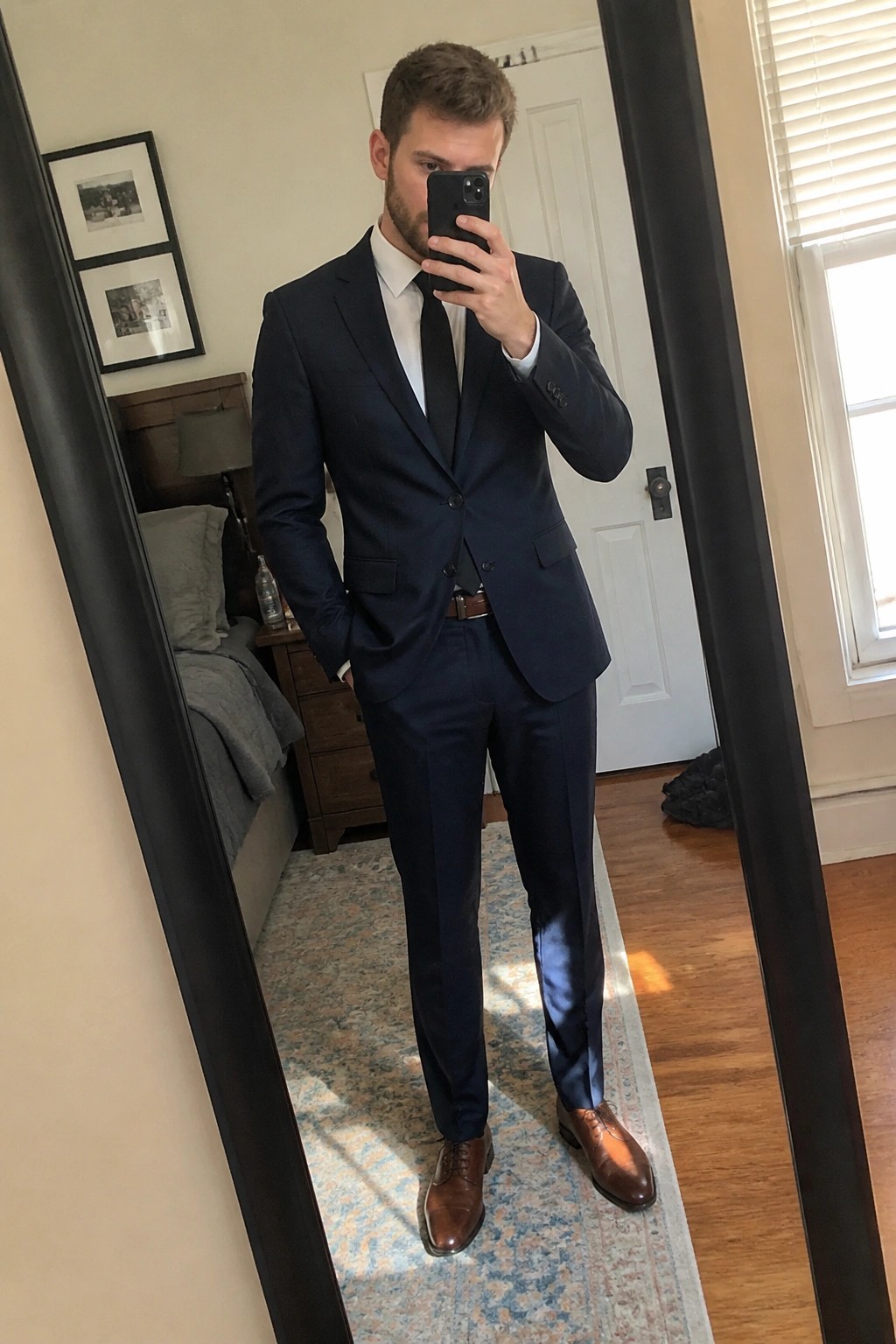 Man in navy suit and black tie taking mirror selfie in bedroom.
