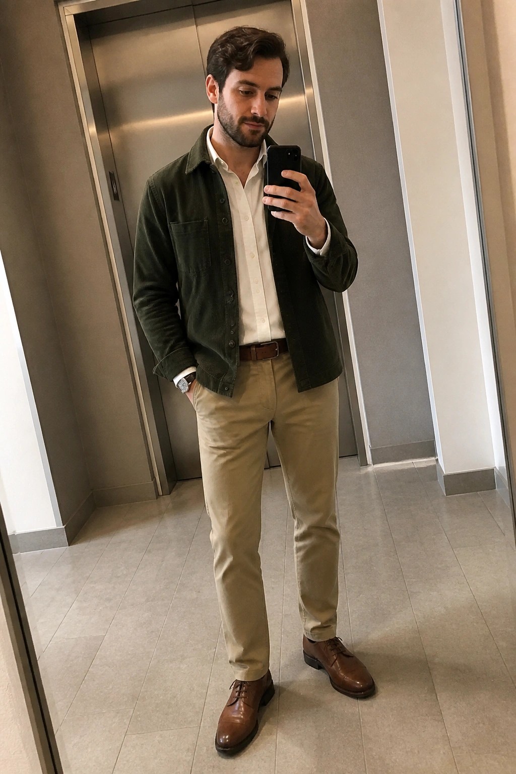 Man standing in an elevator mirror selfie wearing a dark green corduroy shirt jacket open over a white button down shirt tucked into khaki pants with a brown leather belt and brown leather dress shoes.