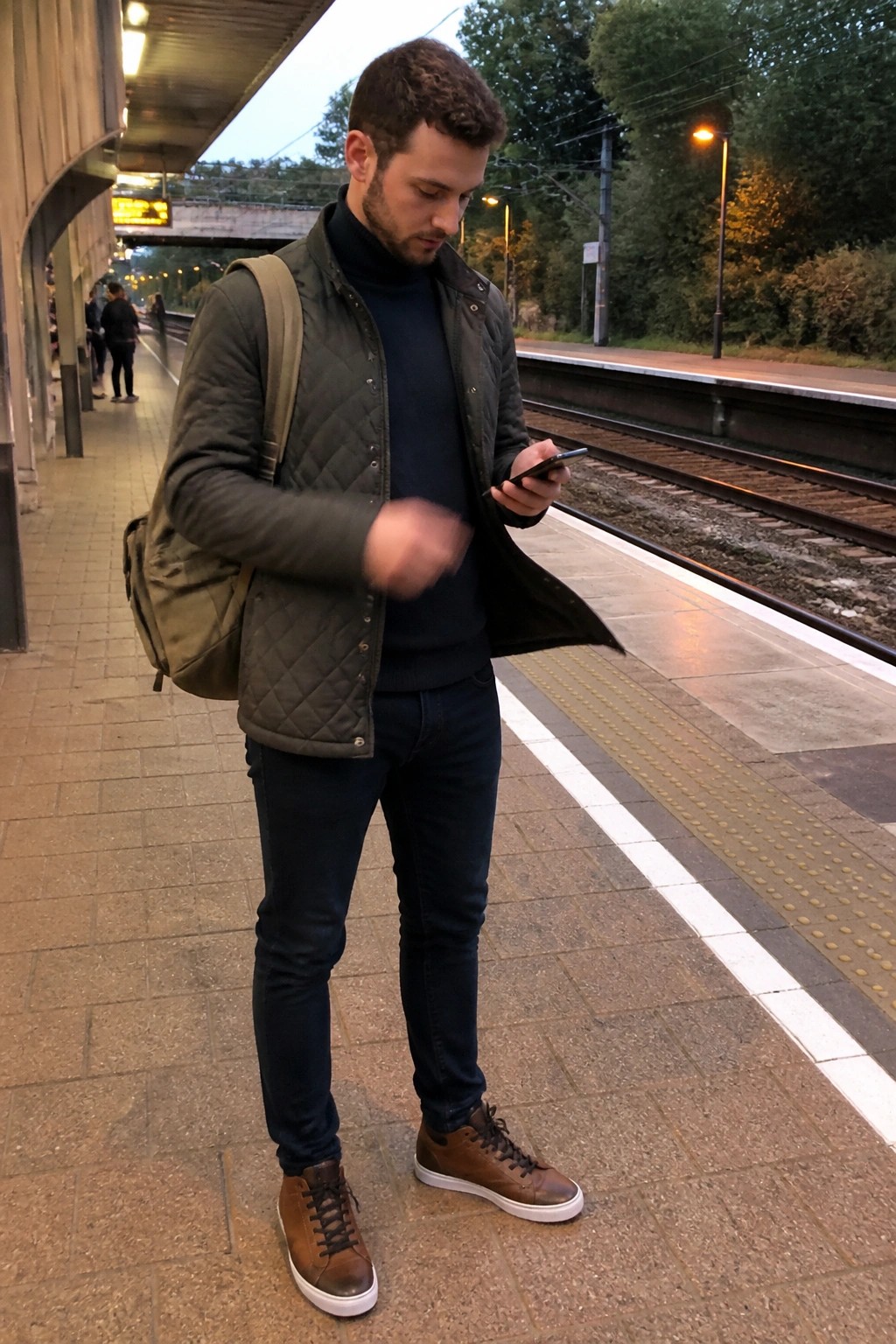 Man in green quilted vest over navy sweater checks phone on train platform.