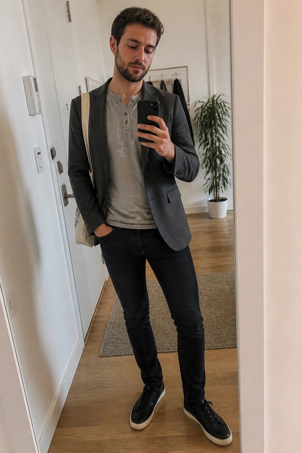 Man in gray blazer, henley shirt, dark jeans takes mirror selfie with tote bag