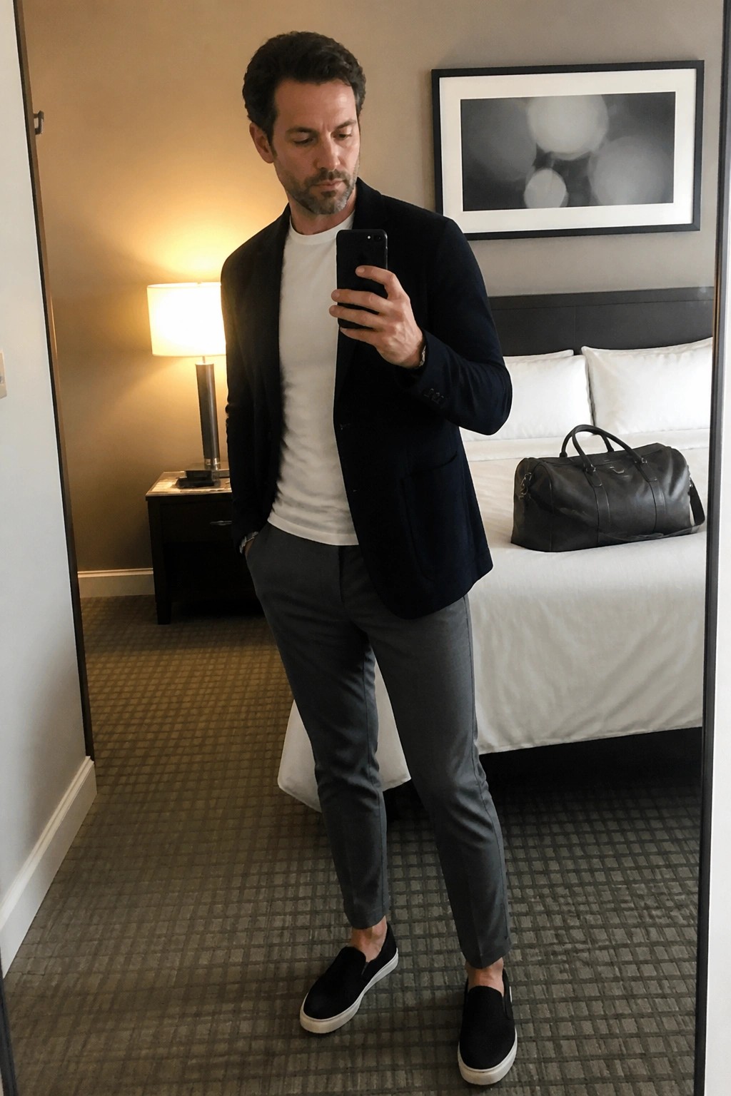 A man taking a mirror selfie wearing a navy blazer over a white t-shirt with gray trousers and black slip-on sneakers.