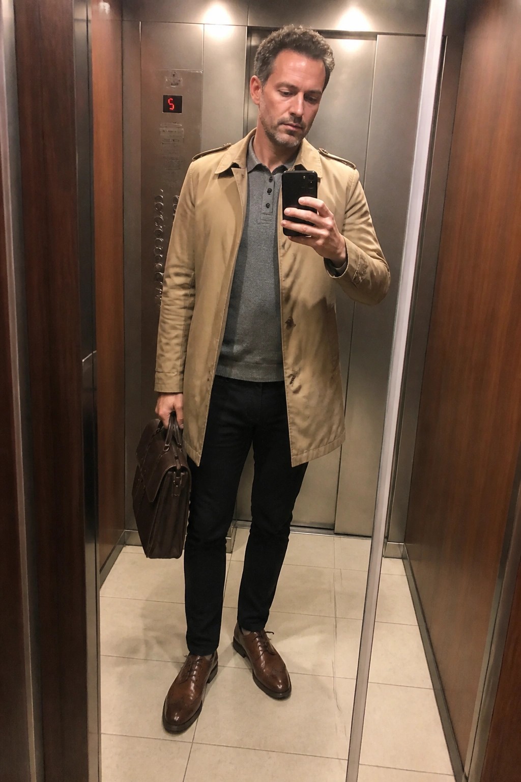 Man standing in an elevator wearing a beige trench coat over a gray polo sweater with black pants, brown leather shoes, and holding a brown leather bag while checking his phone.