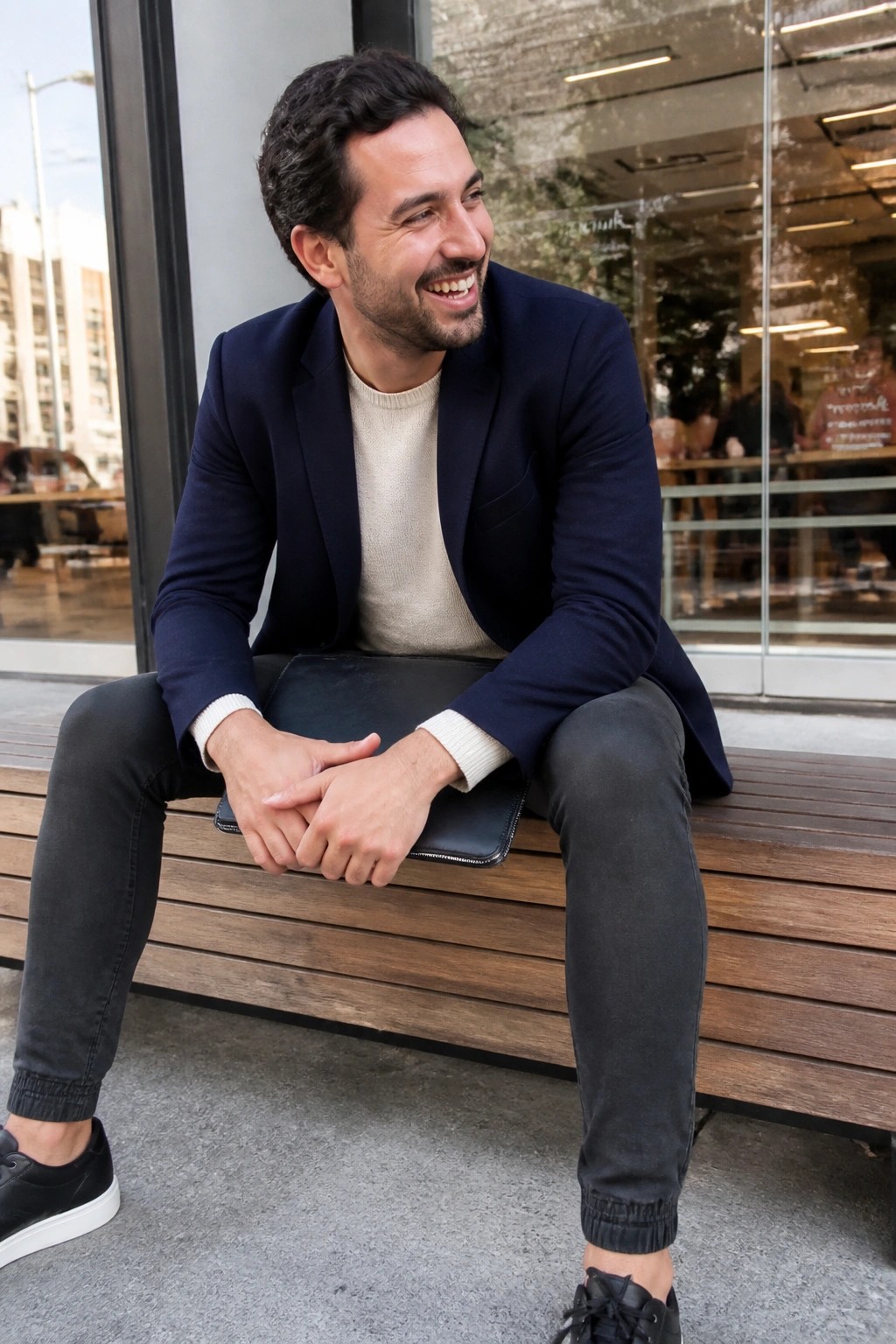 Smiling man in navy blazer and beige sweater holds black leather pouch on bench