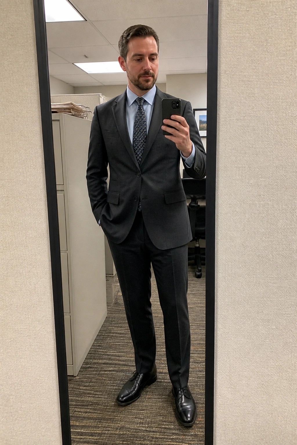 Man in dark gray suit takes mirror selfie in office with phone