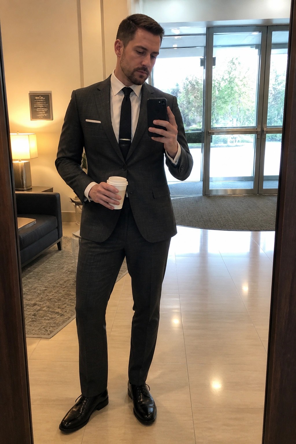 Man in gray plaid suit taking mirror selfie with coffee and phone.
