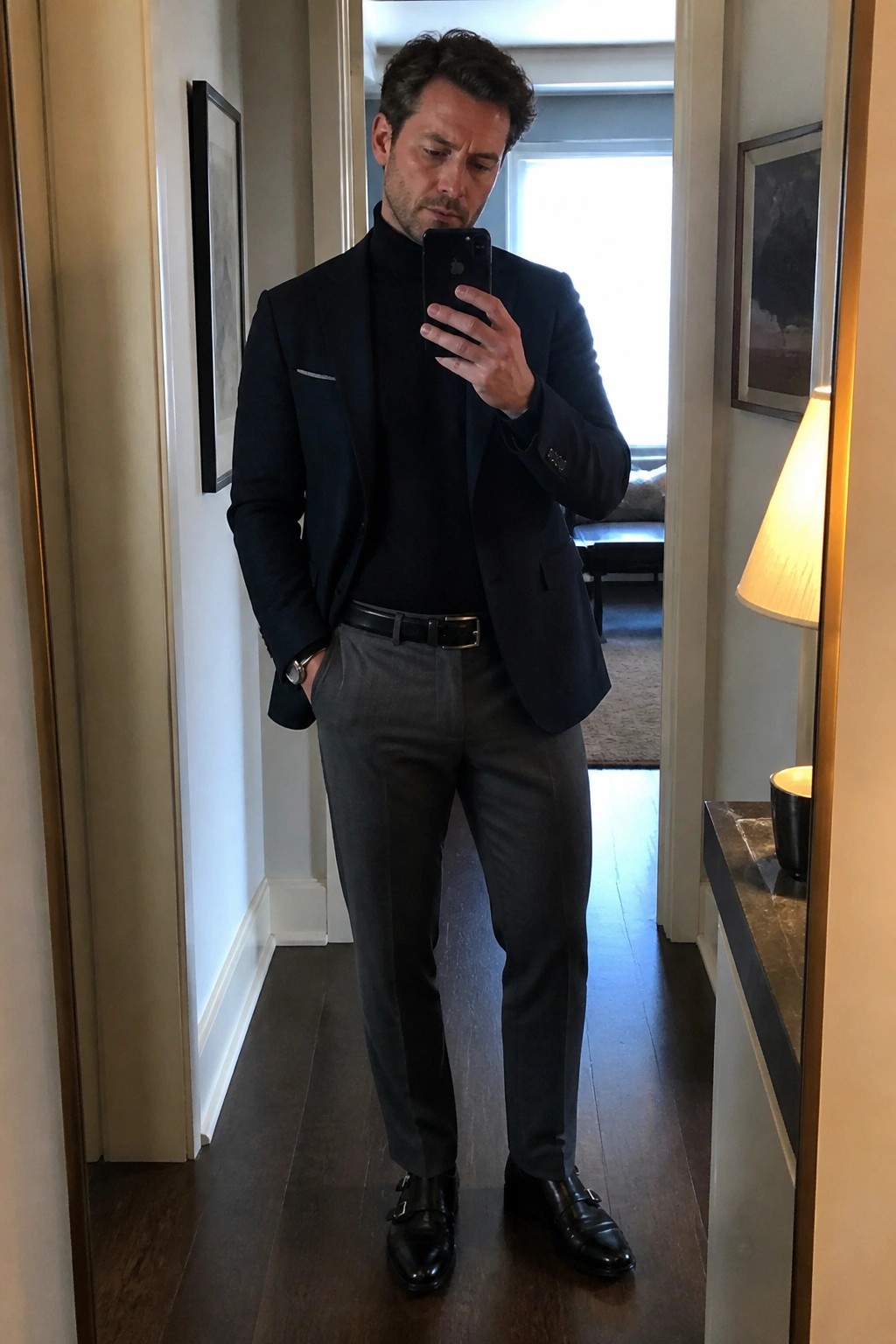 Man in navy blazer over black turtleneck taking mirror selfie in hallway