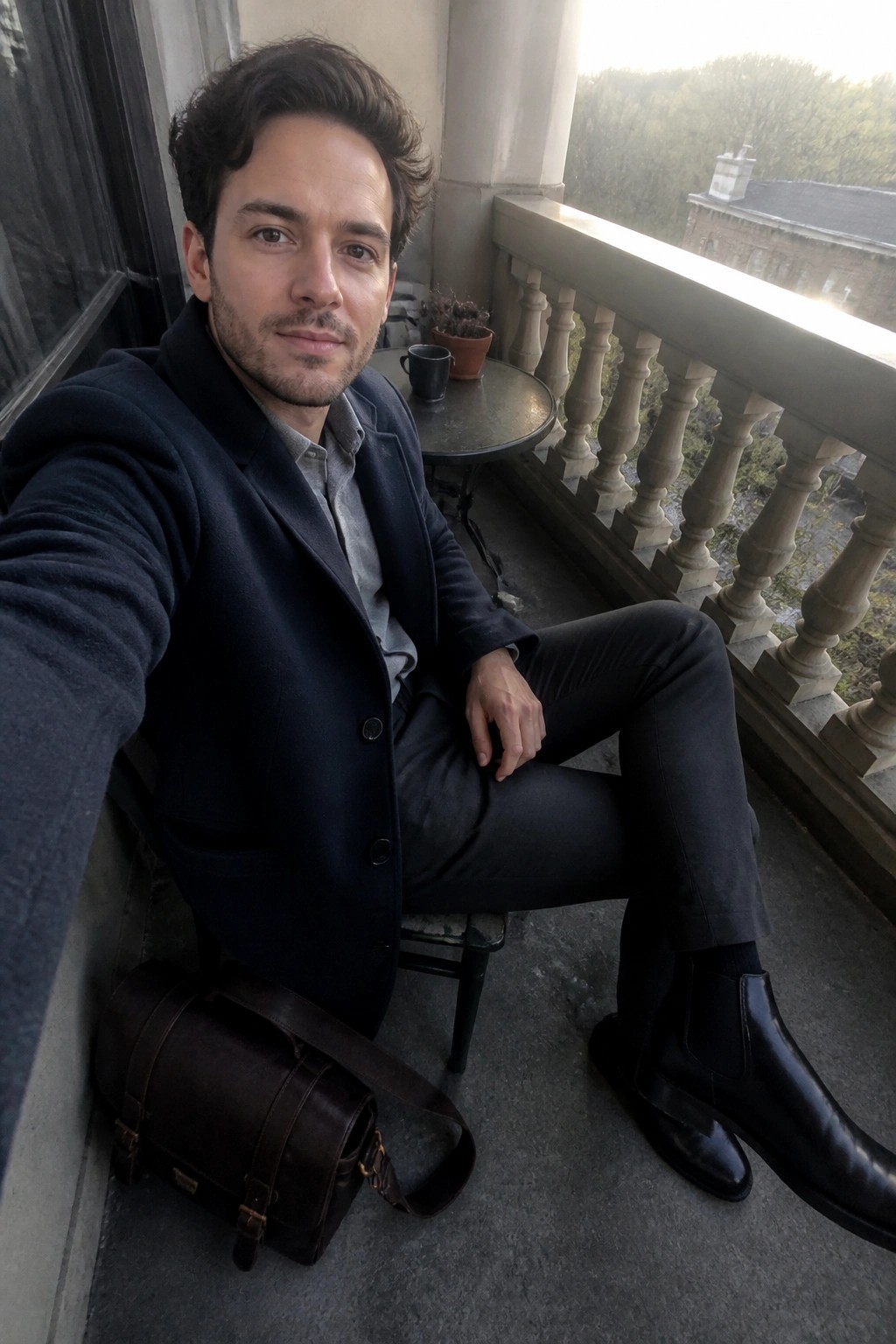 Man in navy overcoat and black boots sits on balcony beside leather bag.