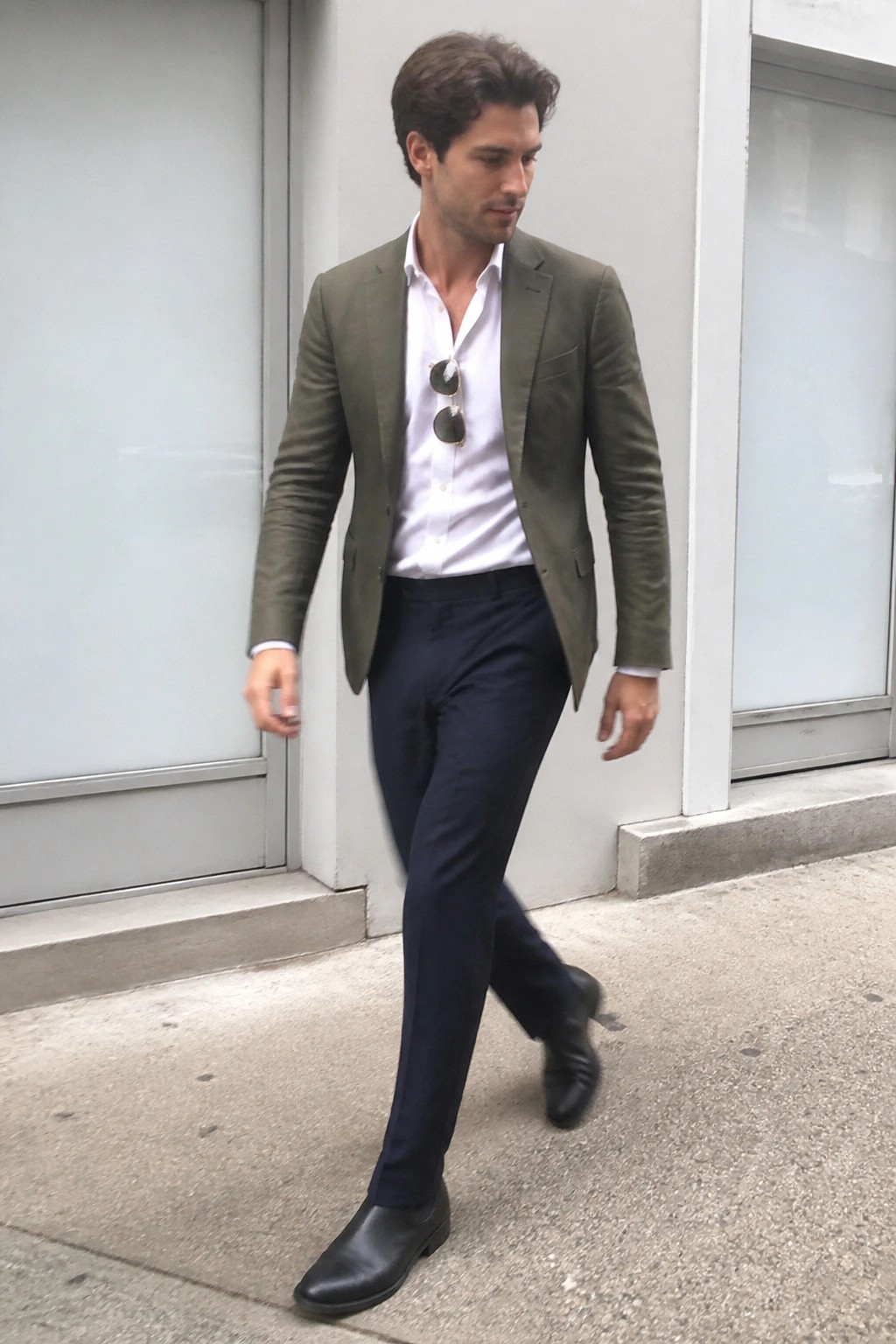 Man in olive blazer, white shirt, and navy pants walks outdoors with sunglasses tucked in collar