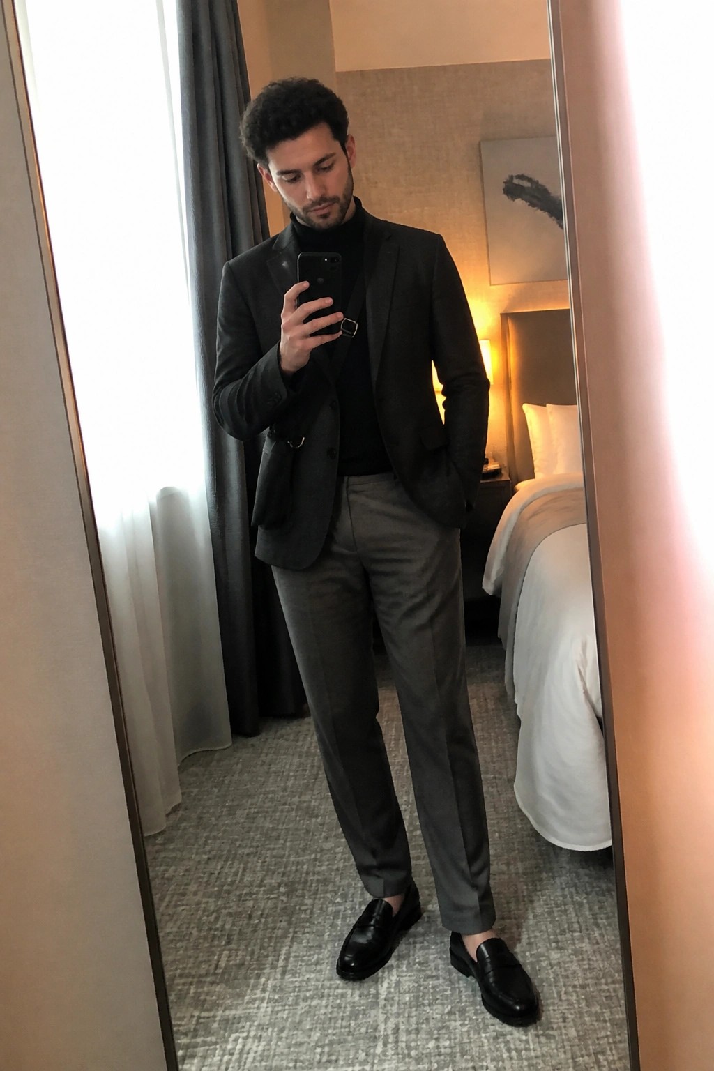 Man in dark blazer over black turtleneck and gray pants taking mirror selfie in hotel room