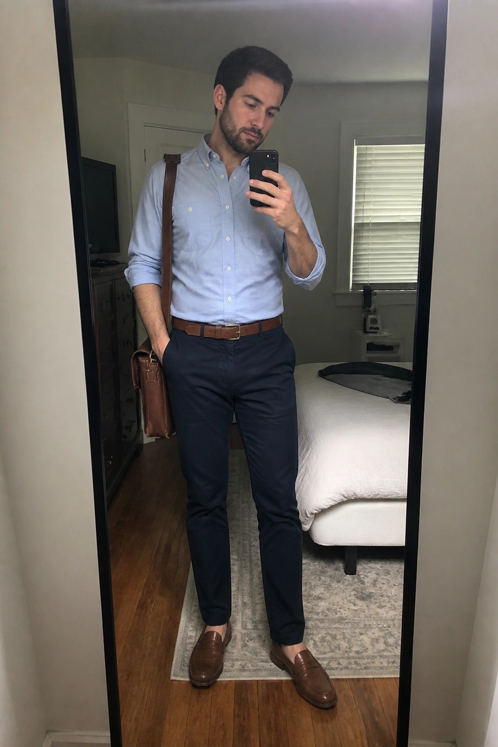 Man in light blue shirt and navy pants taking mirror selfie in bedroom