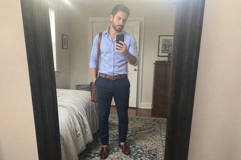 Man in light blue shirt and navy pants taking mirror selfie in bedroom