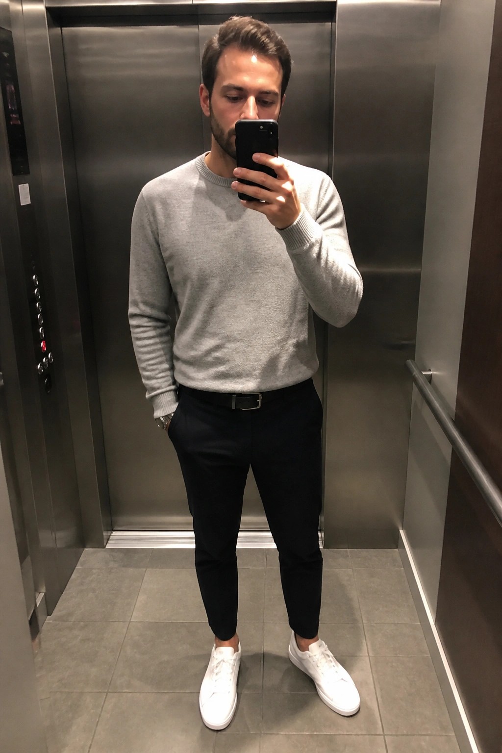 Man in gray sweater and black pants taking mirror selfie in elevator wearing white sneakers
