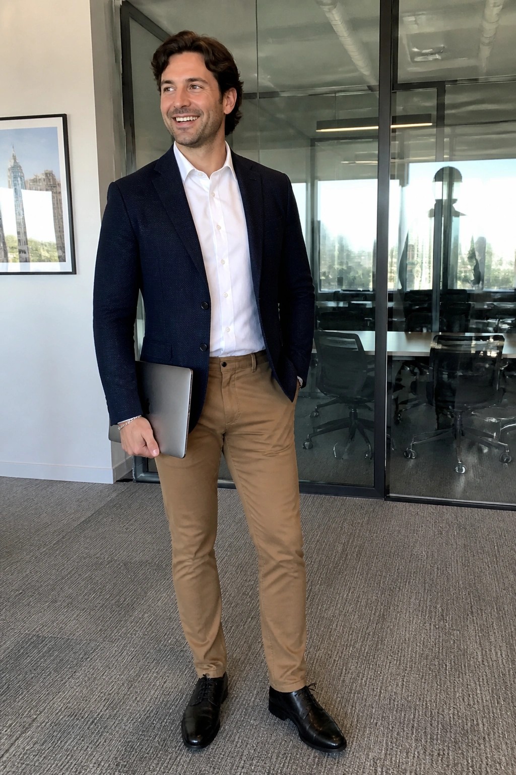 Smiling man in navy blazer and khaki pants holds laptop in modern office