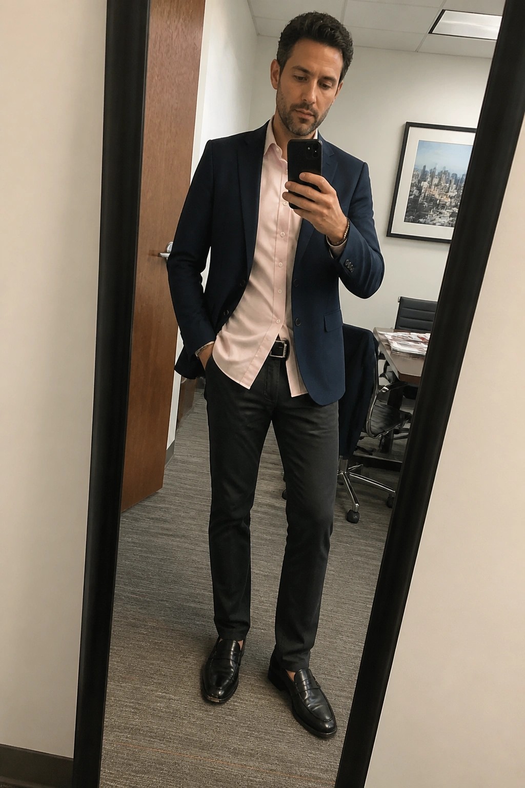 Man in navy blazer and pink shirt taking mirror selfie in office