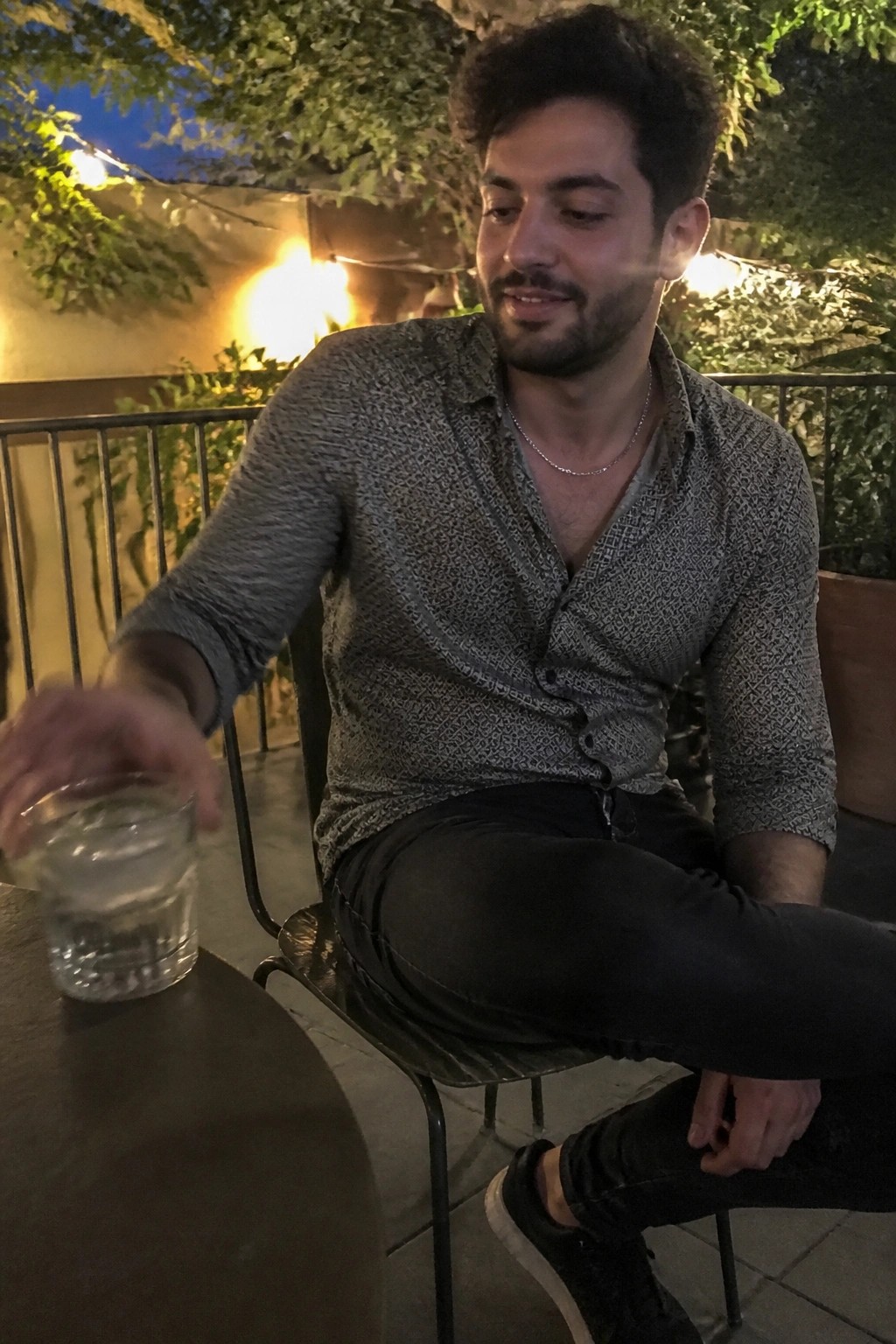Bearded man in patterned shirt sits at night table, hand on water glass.