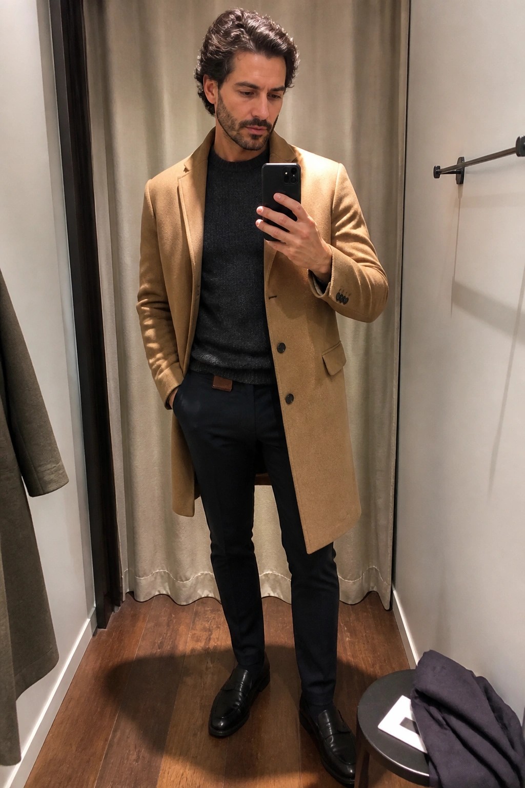 Man in tan overcoat over gray sweater taking mirror selfie in fitting room