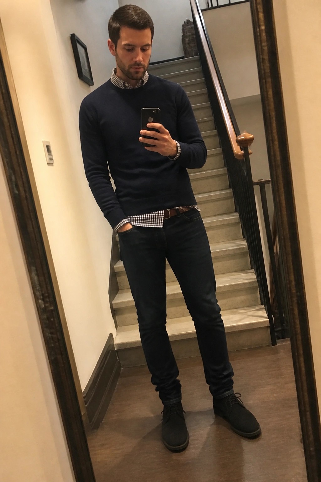 Man wearing navy crewneck sweater over blue and white check shirt tucked into dark slim jeans with brown belt and black suede chukka boots while holding a phone for a mirror selfie in a hallway with stairs behind him.