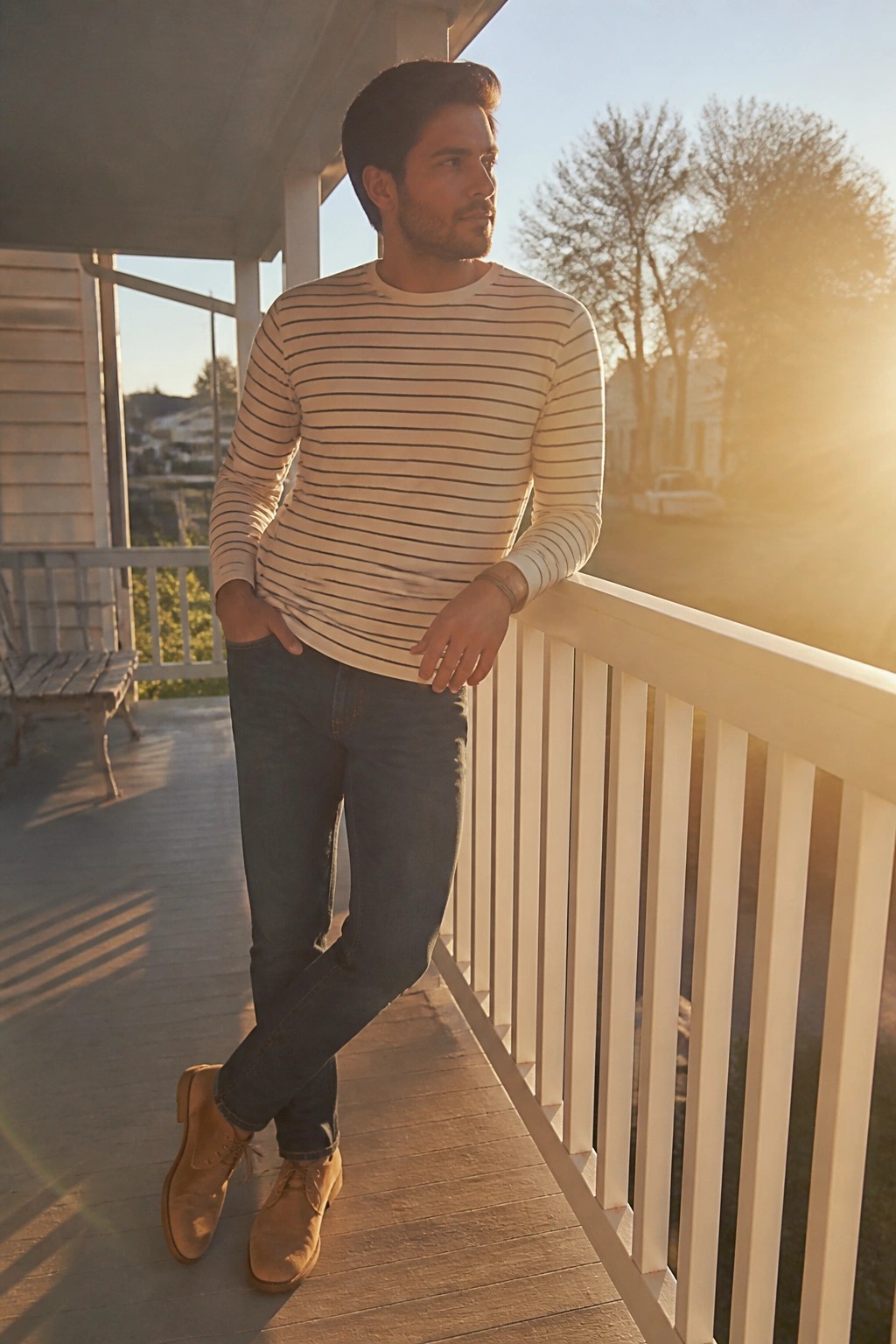 Man in striped shirt and jeans leaning on sunny porch railing at sunset