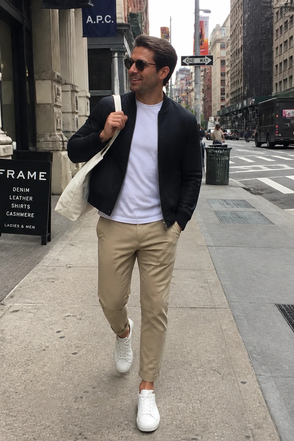 Man in black bomber jacket, white tee, khaki pants, white sneakers with tote bag on sidewalk