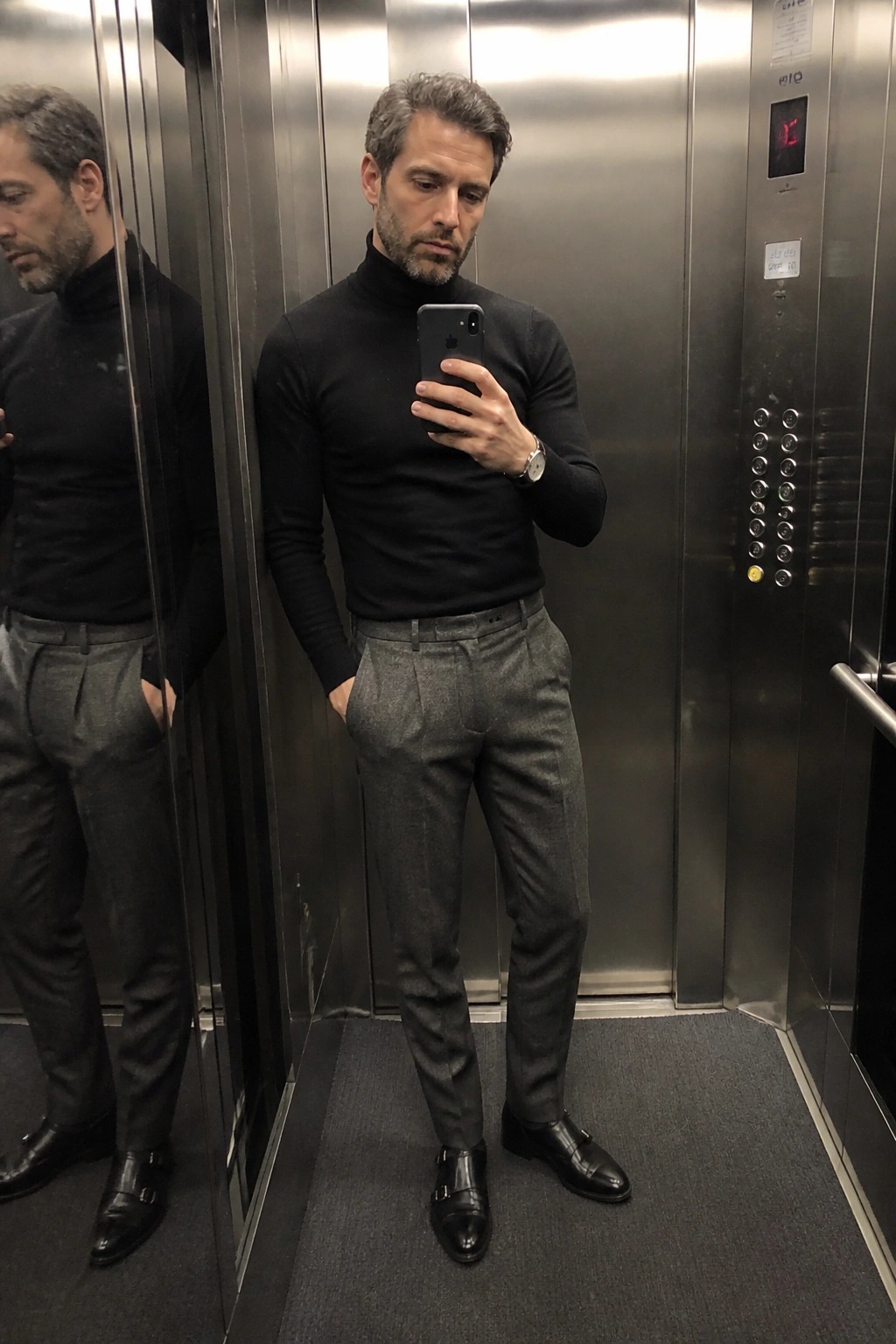 A man in a fitted black turtleneck sweater tucked into gray pleated wool trousers with black double monk strap shoes standing in an elevator while holding a phone for a mirror selfie.