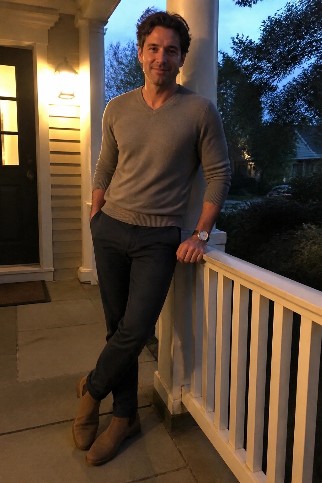 Man in gray V-neck sweater leans on white porch railing at dusk