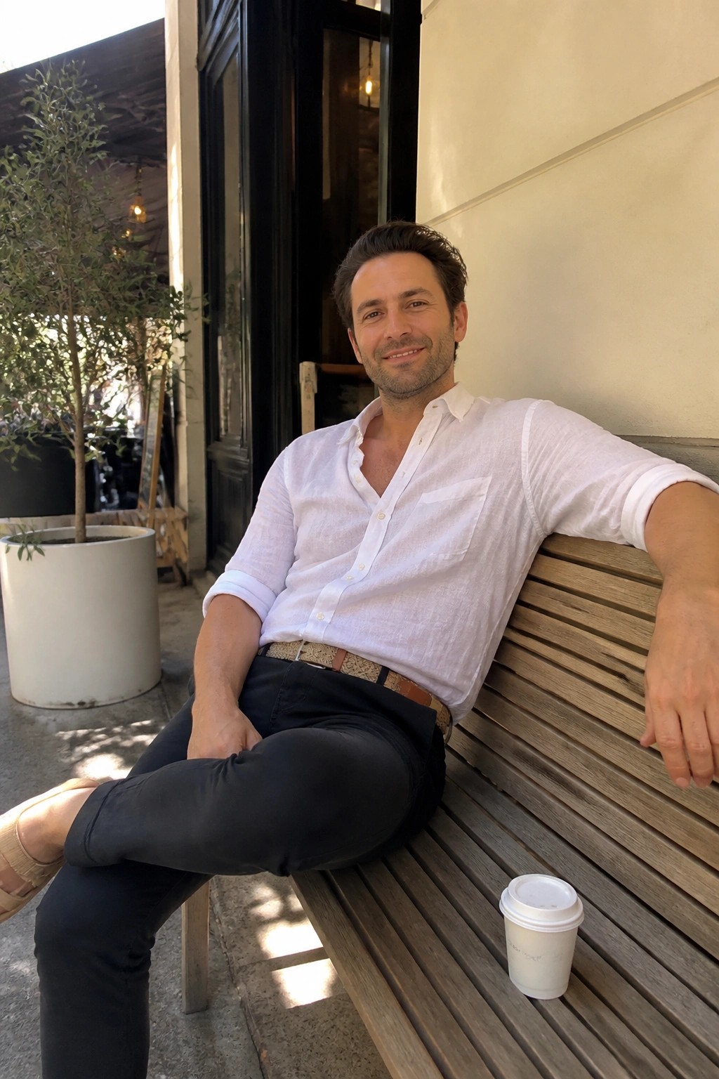 Man in white shirt and dark pants relaxes on wooden bench beside coffee cup