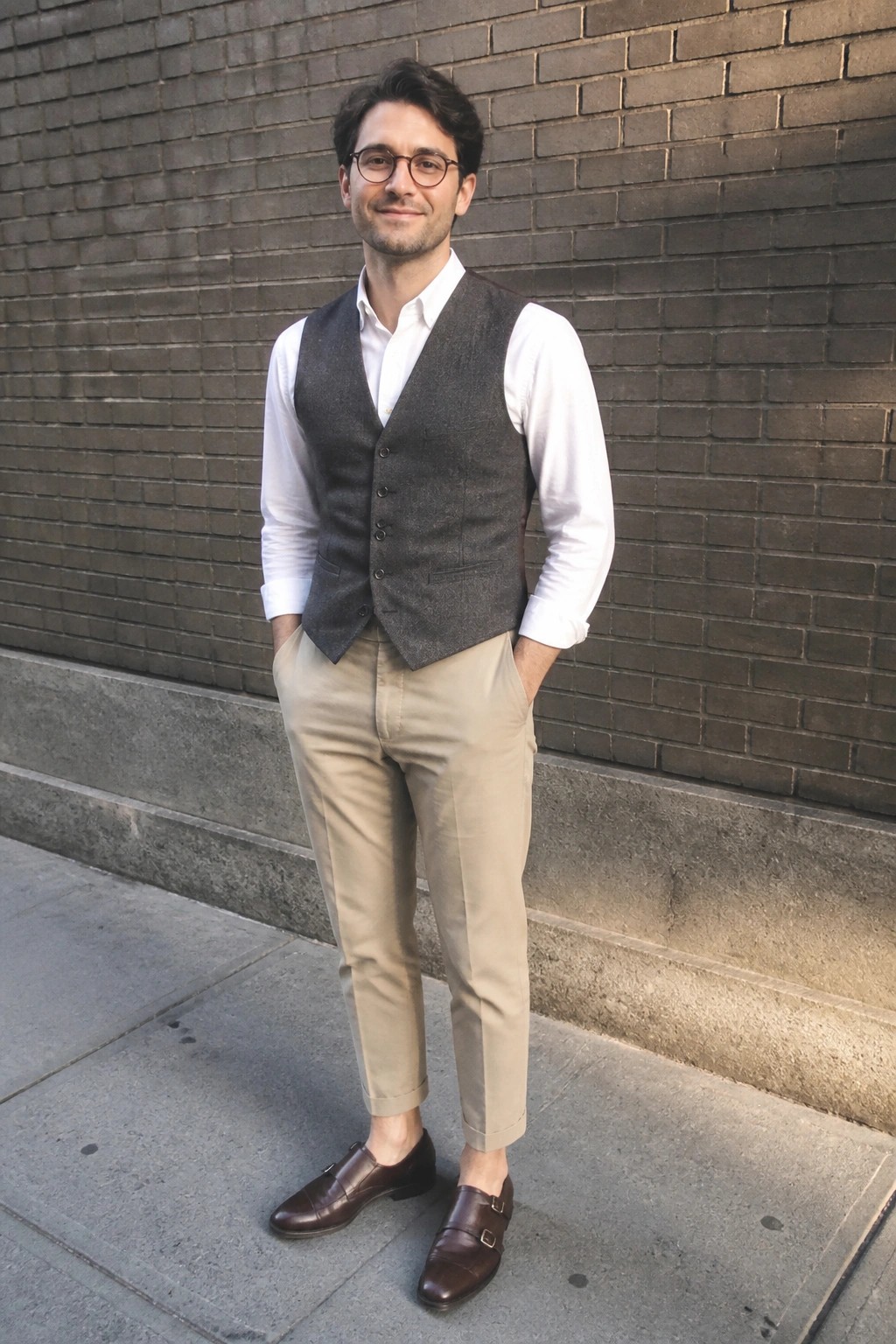 Man in white shirt, gray vest, beige pants and brown monk shoes by brick wall