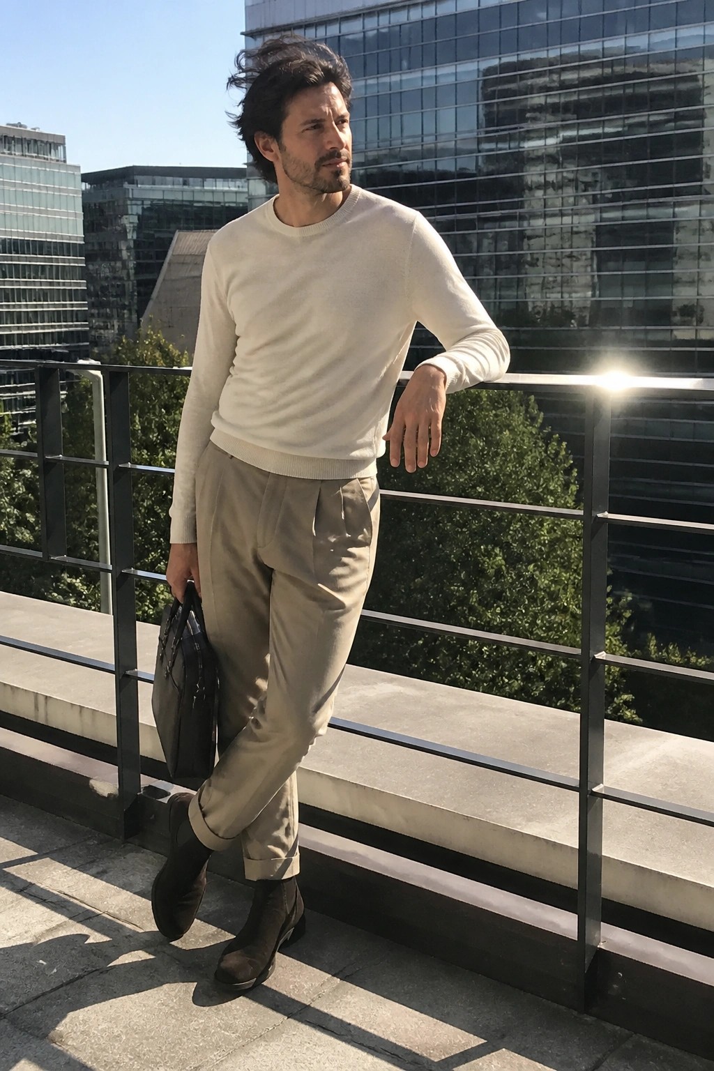 Man in cream sweater and beige pants leaning on railing holding leather bag