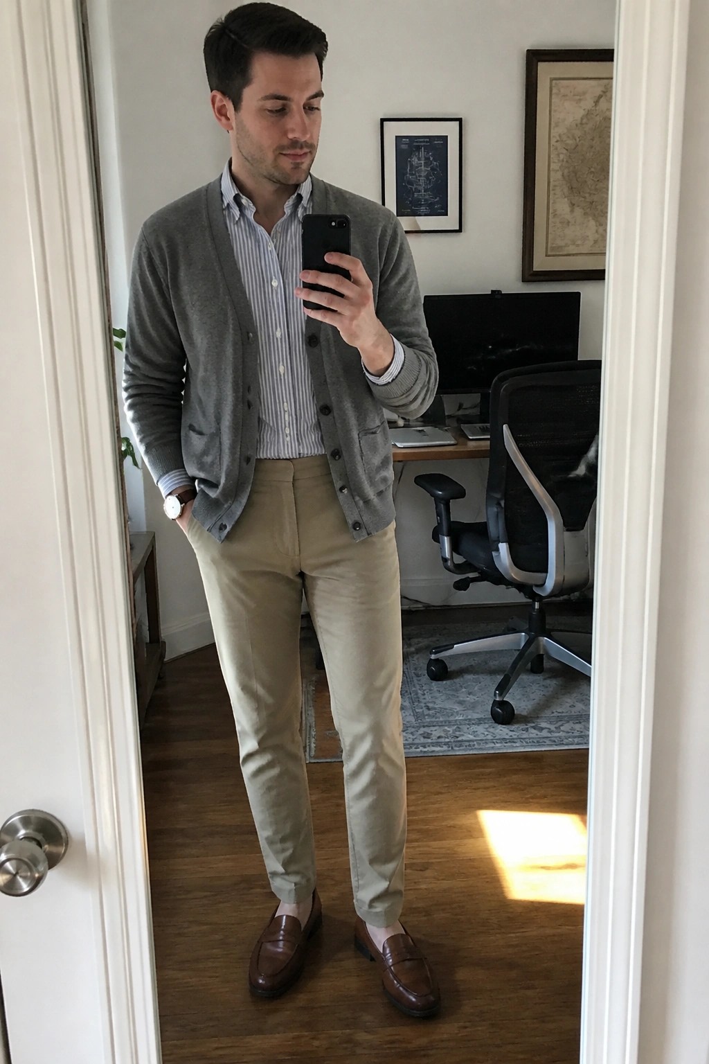 Man in gray cardigan over striped shirt and khaki pants taking mirror selfie