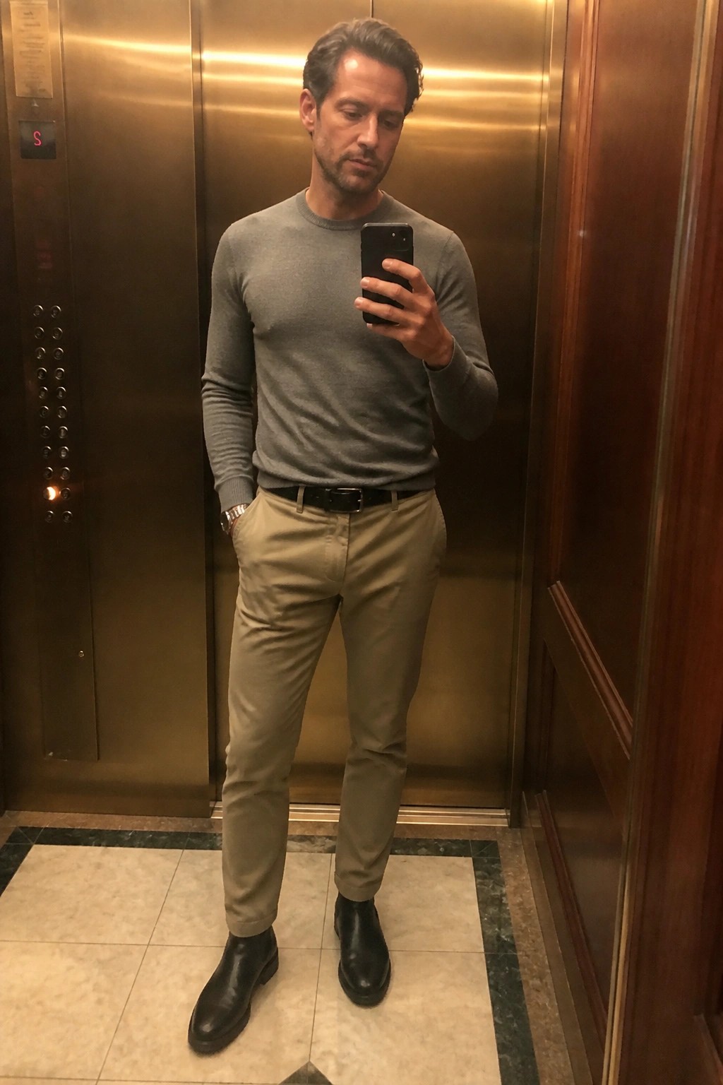 Man in gray sweater and khaki pants holding phone in elevator