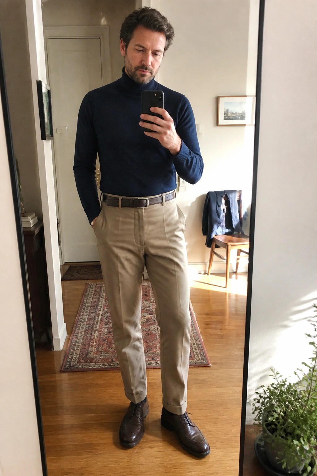 Man wearing a navy turtleneck sweater tucked into light khaki trousers with a brown leather belt and brown leather brogue shoes.
