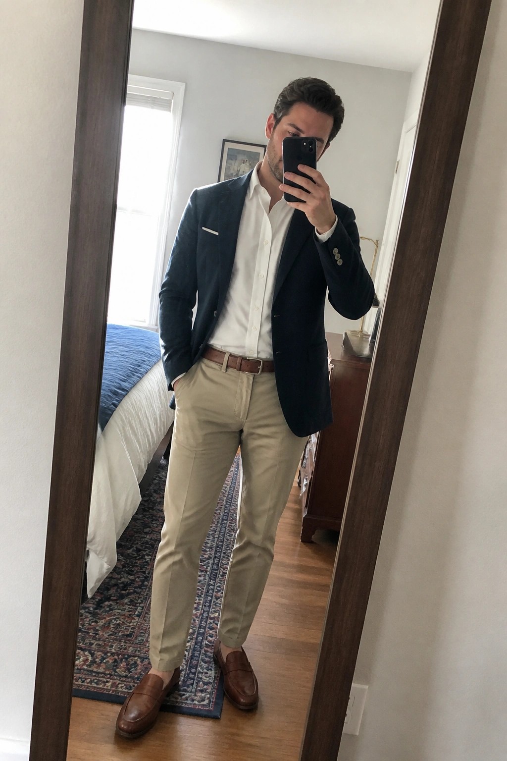 Man in navy blazer, white shirt, and khakis taking bedroom mirror selfie