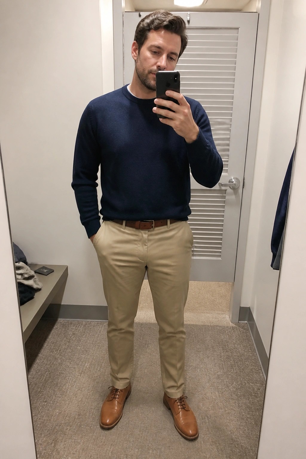 Man in navy sweater and khaki pants taking mirror selfie with phone