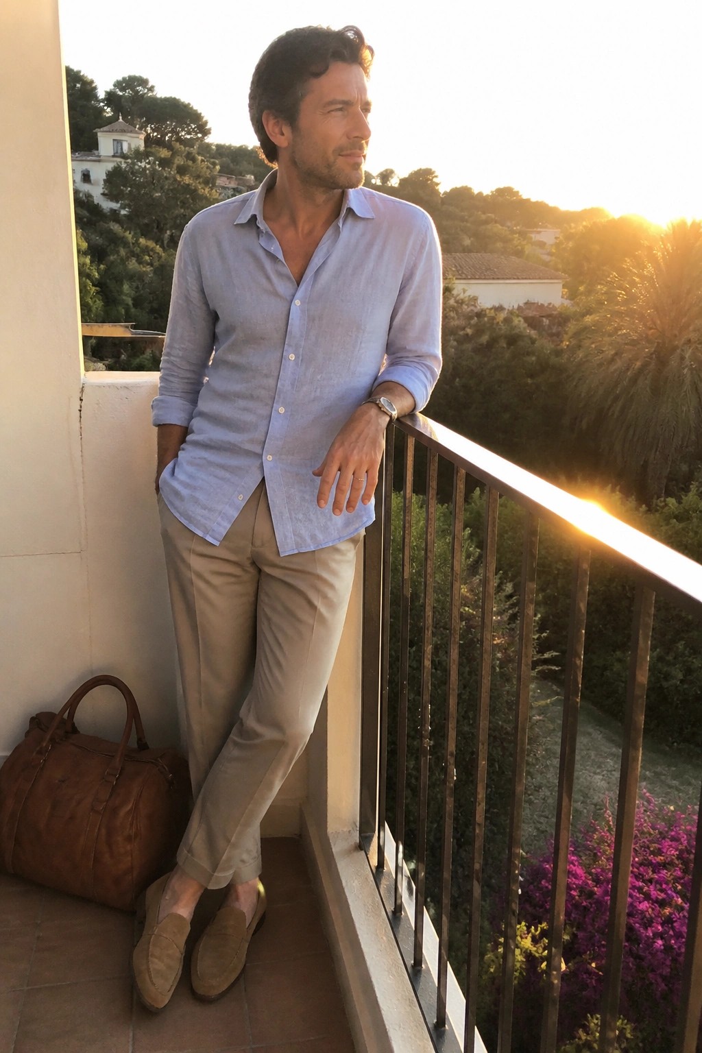 Man in light blue shirt and beige pants leaning on balcony railing at sunset