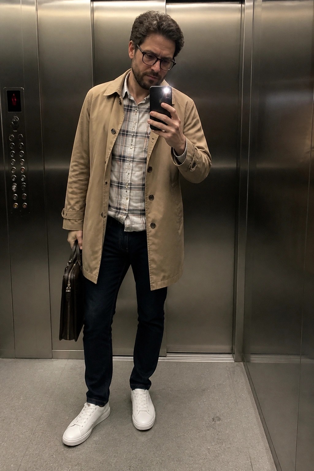 Man in tan trench coat over plaid shirt holds phone and bag in elevator