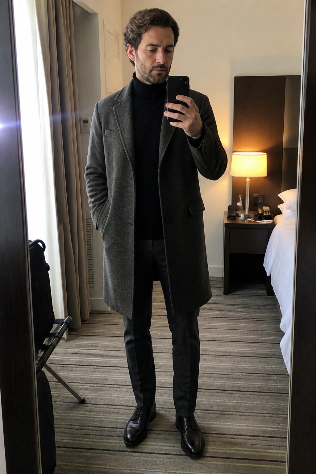 Man in gray overcoat and black turtleneck taking mirror selfie in hotel room
