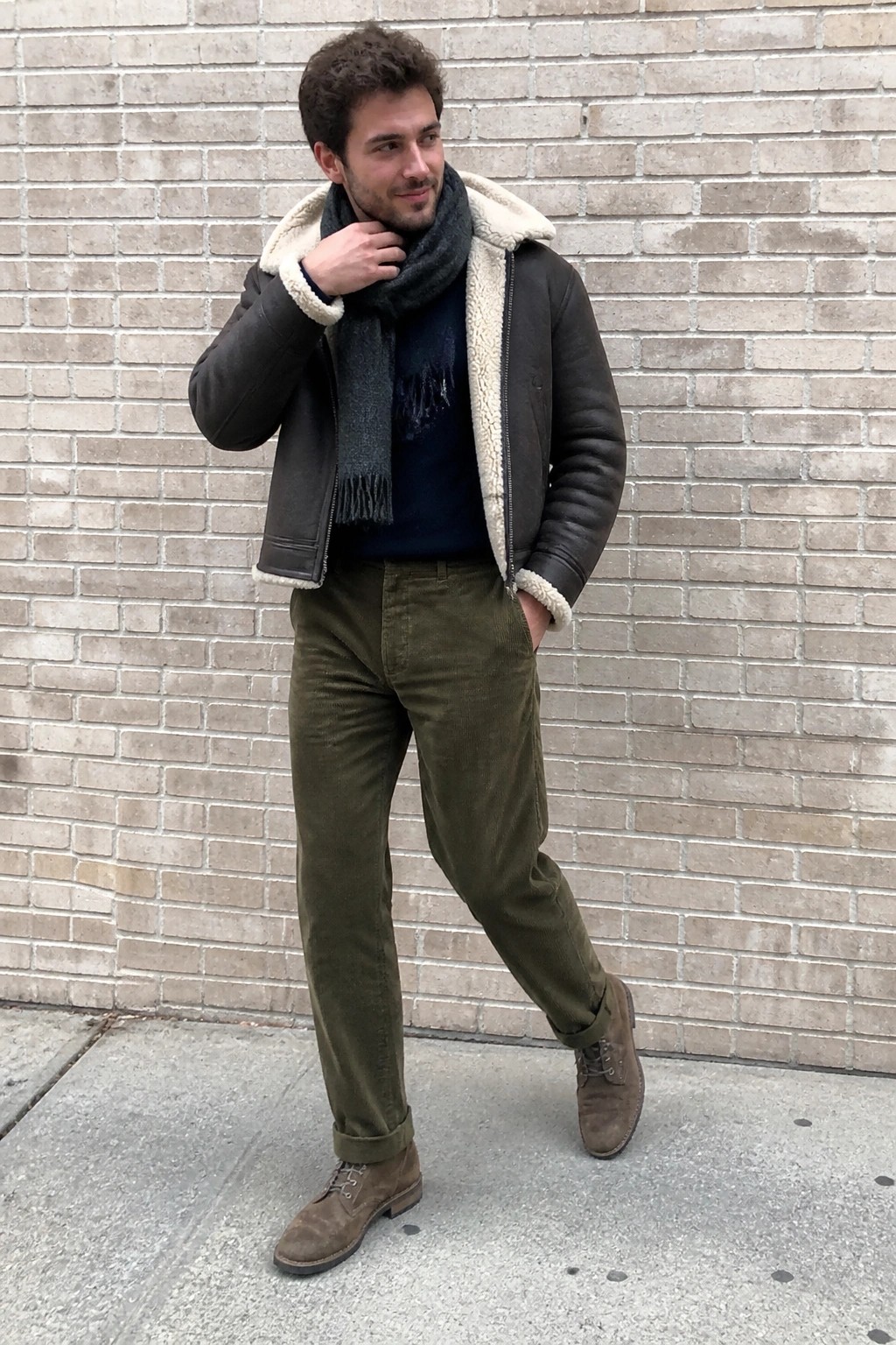 Man in brown shearling jacket, scarf, and olive corduroy pants by brick wall