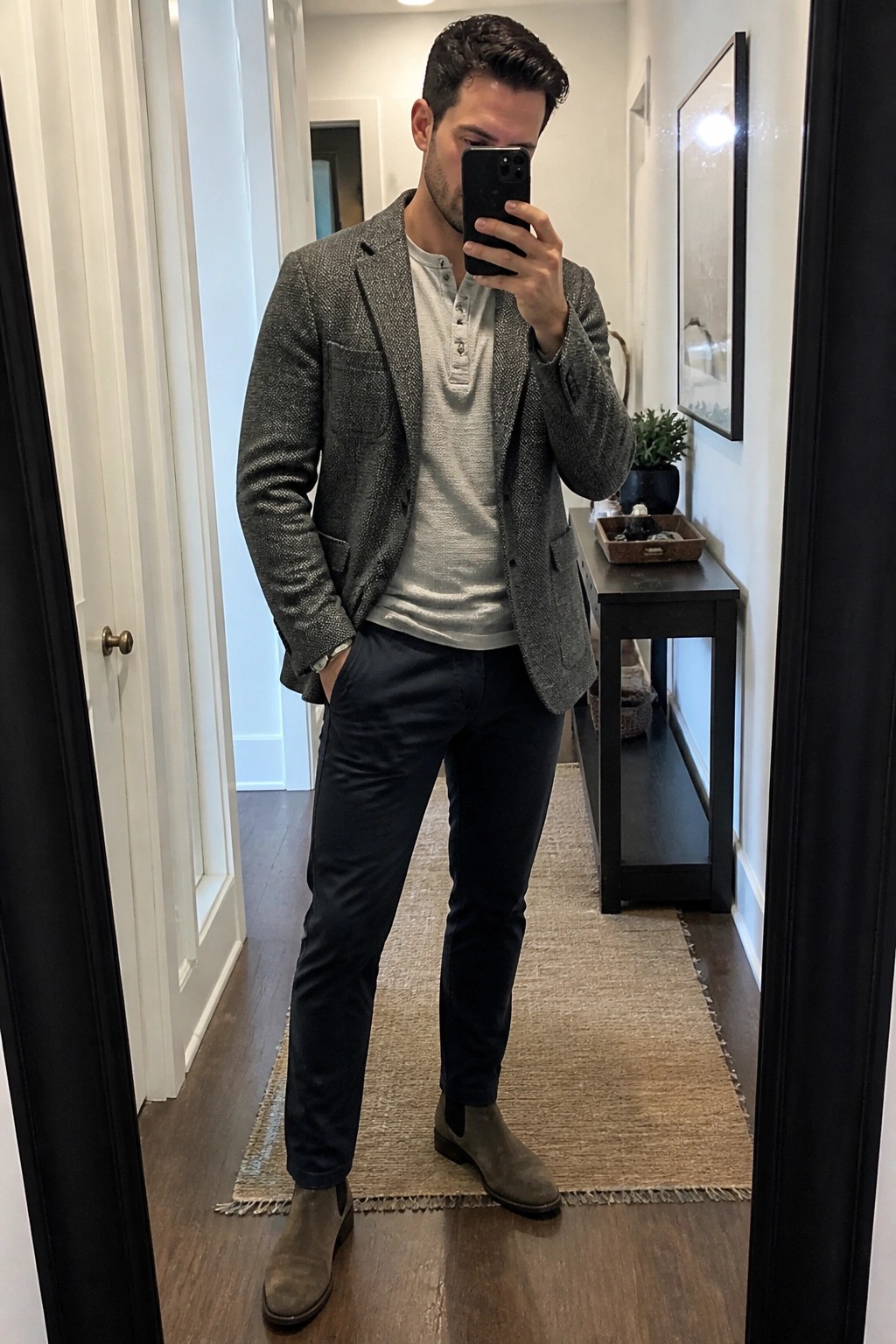 Man in gray tweed blazer, henley shirt, and suede boots taking mirror selfie