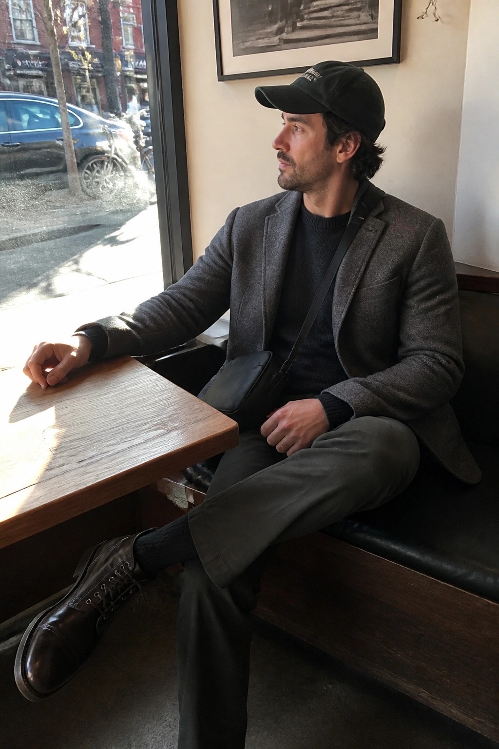 Man in gray blazer and black cap sits by sunny cafe window with crossbody bag