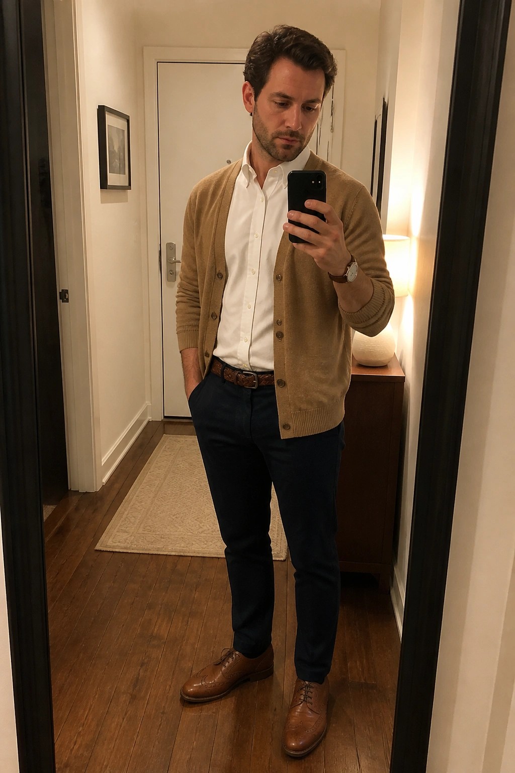 A man in a white button down shirt layered under a tan cardigan with navy trousers a brown woven belt and brown brogue shoes holding a phone for a mirror selfie.