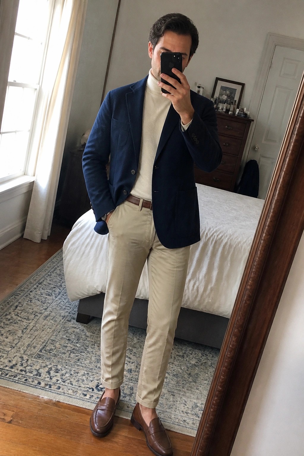 Man in navy blazer over cream turtleneck with khaki pants taking mirror selfie