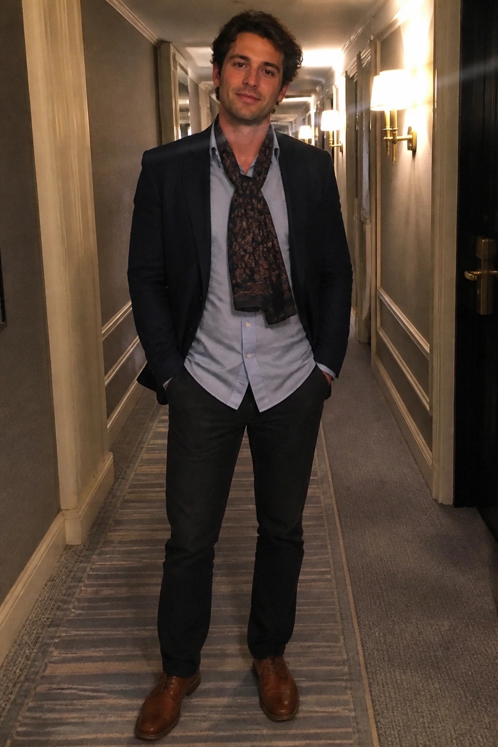 Man in navy blazer and patterned scarf stands in elegant carpeted hallway