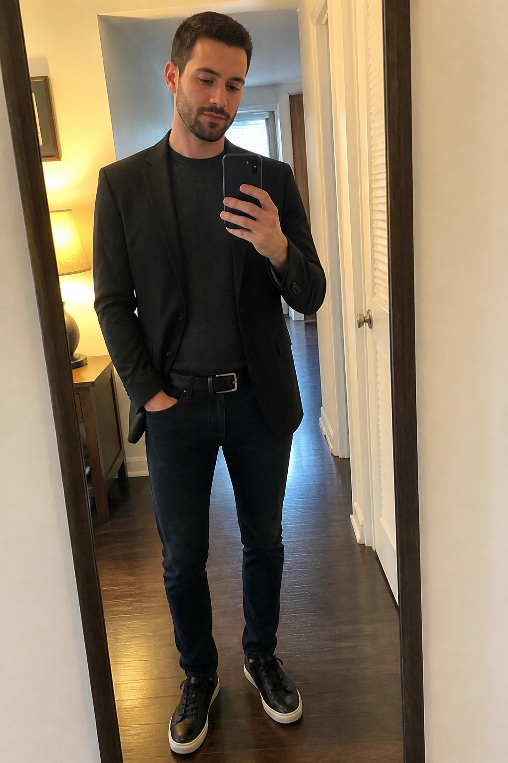 Man in black blazer and jeans taking mirror selfie in hallway.